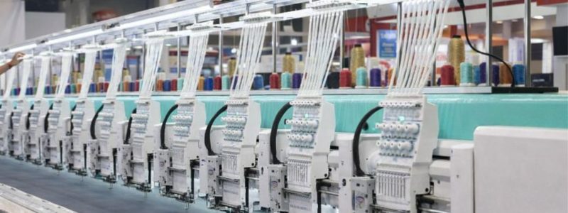 Rad av industriella broderimaskiner med trådar i flera färger, redo för tryck på tyg.